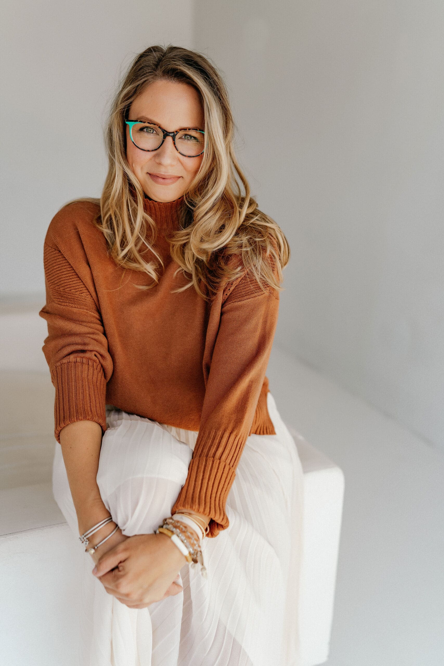 Professional wellness practitioner in cozy rust-colored sweater and glasses, sitting comfortably in white clothing against a neutral background, embodying a warm and approachable healthcare approach