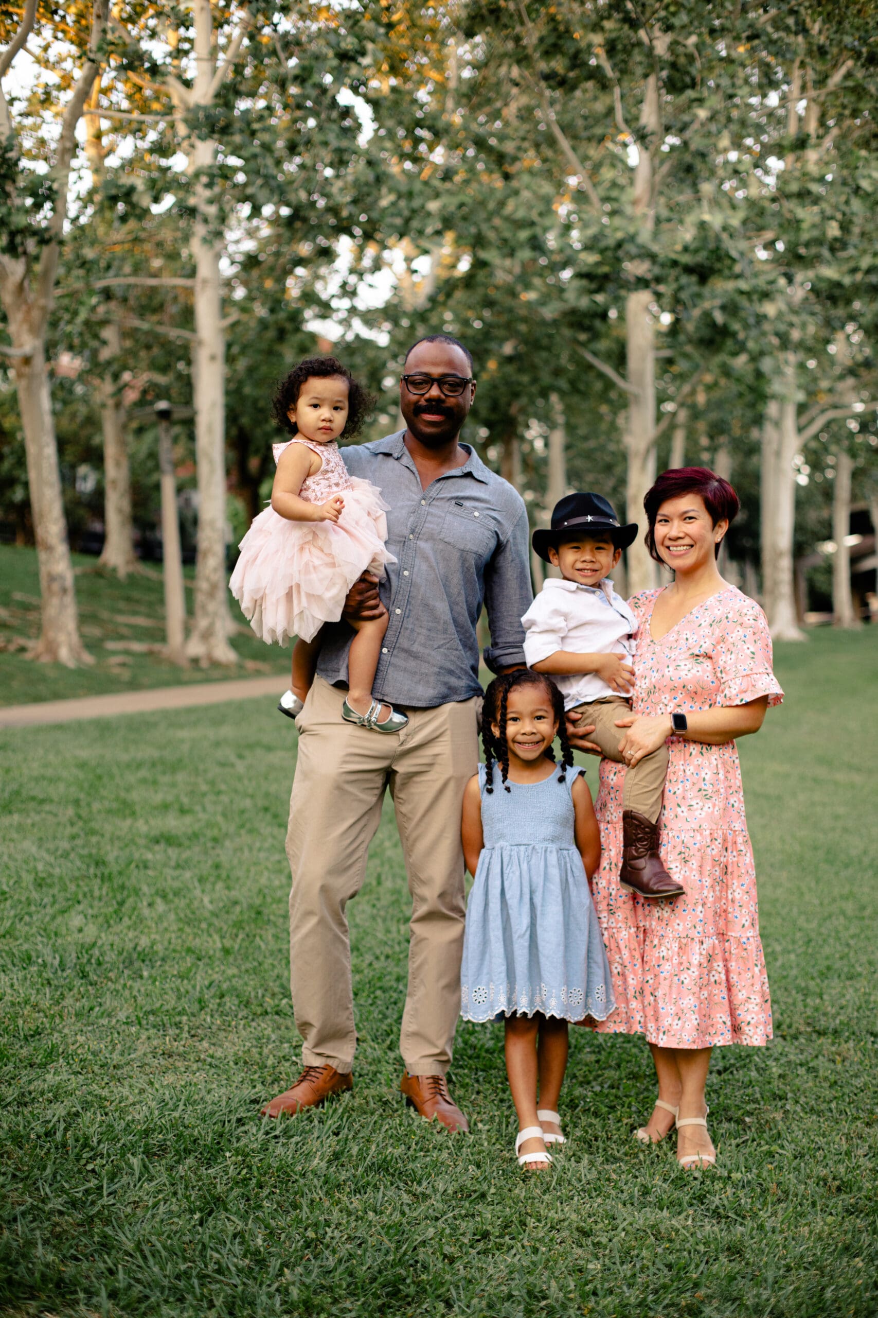 The Woodlands family photographer captures a beautiful family of five posing on green grass with trees in the background, featuring parents and three children in coordinated pastels